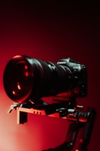 A professional camera mounted on a rig or stabilizer, highlighted by dramatic red lighting. The background is dark, accentuating the sleek design of the camera and its prominent lens.