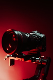 A professional camera mounted on a rig or stabilizer, highlighted by dramatic red lighting. The background is dark, accentuating the sleek design of the camera and its prominent lens.