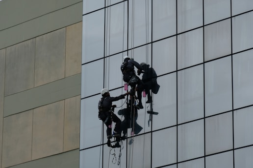 Two workers wearing harnesses and helmets are suspended on ropes while cleaning the glass windows of a tall building. Their equipment includes buckets and squeegees.