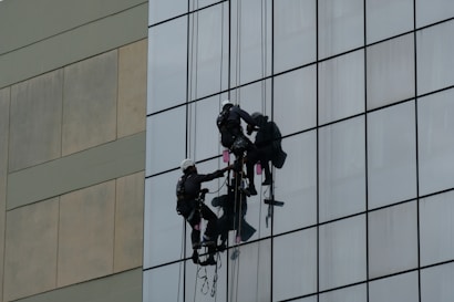 Two workers wearing harnesses and helmets are suspended on ropes while cleaning the glass windows of a tall building. Their equipment includes buckets and squeegees.