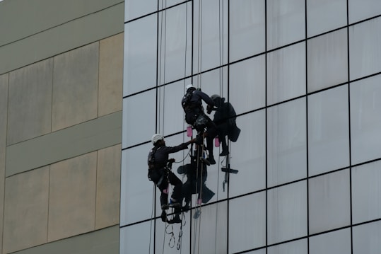 Two workers wearing harnesses and helmets are suspended on ropes while cleaning the glass windows of a tall building. Their equipment includes buckets and squeegees.