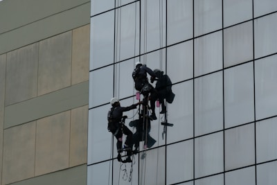 Two workers wearing harnesses and helmets are suspended on ropes while cleaning the glass windows of a tall building. Their equipment includes buckets and squeegees.