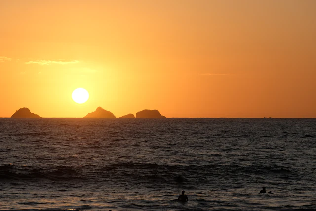 The sun setting over the ocean off Arpoador, with the Cagarras Islands silhouetted on the horizon and surfers in the water.