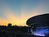 An outdoor concert venue filled with a large audience enjoying a live performance on a stage. The sky is a gradient of blue and orange, indicating a sunset or sunrise, creating a picturesque backdrop. A modern architectural structure provides coverage over the stage, which is lit with various lights and decorations.