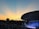 An outdoor concert venue filled with a large audience enjoying a live performance on a stage. The sky is a gradient of blue and orange, indicating a sunset or sunrise, creating a picturesque backdrop. A modern architectural structure provides coverage over the stage, which is lit with various lights and decorations.