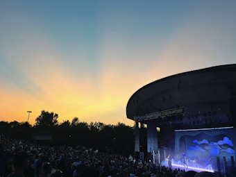 An outdoor concert venue filled with a large audience enjoying a live performance on a stage. The sky is a gradient of blue and orange, indicating a sunset or sunrise, creating a picturesque backdrop. A modern architectural structure provides coverage over the stage, which is lit with various lights and decorations.