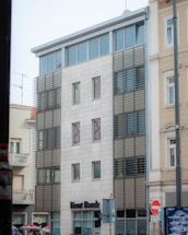 A modern building with a light facade and horizontal sunshades covering the windows. The structure is adjacent to older, more traditional buildings. Signs of urban elements like a road sign and some advertisements can be seen. The ground level features a bank's signage.