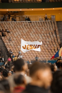 A large crowd of people is gathered in an indoor arena, with rows of empty seats in the background. In the foreground, a flag with the word 'JESUS' is being held up among the crowd. The atmosphere suggests a gathering or event, and another flag with a blue, yellow, and red color scheme is partially visible.
