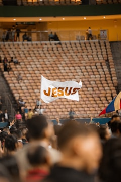 A large crowd of people is gathered in an indoor arena, with rows of empty seats in the background. In the foreground, a flag with the word 'JESUS' is being held up among the crowd. The atmosphere suggests a gathering or event, and another flag with a blue, yellow, and red color scheme is partially visible.