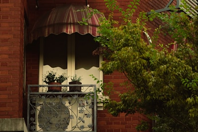 A quaint balcony with a decorative metal railing hosts several small potted plants. An air conditioning unit is partially visible behind the railing. The balcony is shaded by a brown and white striped awning. A brick wall forms the backdrop, with a leafy green tree swaying gently in the foreground.