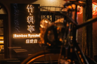 A warmly lit scene features a wooden entrance with soft, glowing signage reading 'Bamboo Ryoutei'. Traditional red lanterns hang above, casting a cozy ambiance. The blurred outline of a bicycle adds depth and a sense of motion to the foreground.