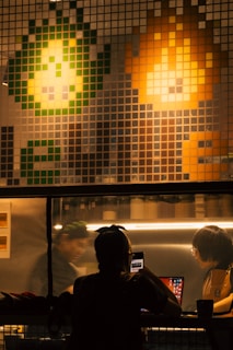 An interior shot of a café or workspace featuring a large pixelated mural on the wall with green and orange abstract shapes resembling characters from a video game. Below the mural, people are engaged in activities, with one person using a smartphone at a counter and another working on a laptop. The lighting creates a warm ambiance.