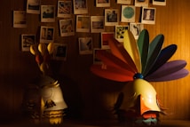 A dimly lit room features a collection of hanging photographs arranged in a grid pattern on a wooden wall. Below the photographs are two artistic sculptures resembling stylized bird heads; one has a multi-colored feather headdress, while the other has a simpler design with a more subtle color palette. The warm light casts soft shadows, creating an intimate atmosphere.