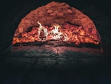 A brick oven interior with flames and glowing embers, highlighting the rustic textures of the stonework. The fire illuminates the inside of the oven, creating a warm and inviting atmosphere.