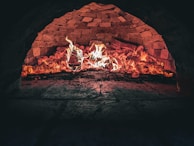 A brick oven interior with flames and glowing embers, highlighting the rustic textures of the stonework. The fire illuminates the inside of the oven, creating a warm and inviting atmosphere.