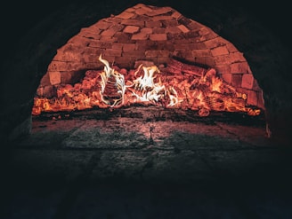 A brick oven interior with flames and glowing embers, highlighting the rustic textures of the stonework. The fire illuminates the inside of the oven, creating a warm and inviting atmosphere.