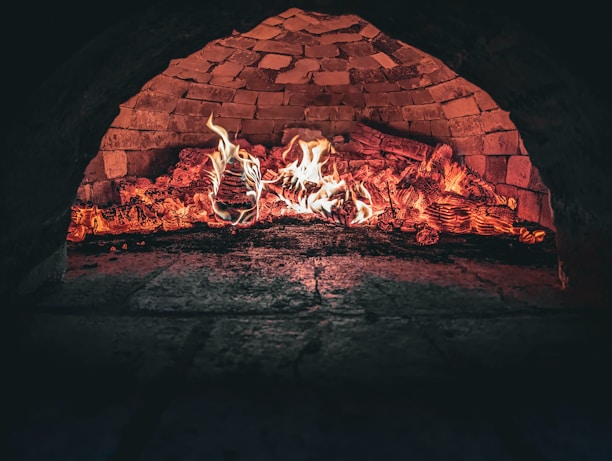 A brick oven interior with flames and glowing embers, highlighting the rustic textures of the stonework. The fire illuminates the inside of the oven, creating a warm and inviting atmosphere.
