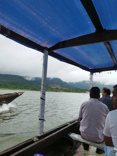 A boat ride with several people seated under a blue tarpaulin cover. The boat is navigating through a river bordered by lush green hills with misty clouds above. A small section of another boat is visible alongside.
