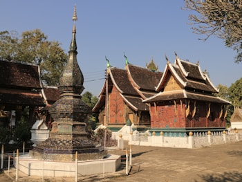 An intricately designed Buddhist temple complex with ornate rooftops and a detailed stupa in the foreground. The structures feature traditional architecture with rich earthy tones and ornate carvings.