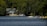 A serene lakeside scene featuring a large, modern house nestled among dense green trees. The calm, blue water reflects the greenery surrounding it. Several boats and docks are visible, including a small motorboat and a yellow watercraft. The setting suggests a peaceful, natural environment.