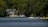 A serene lakeside scene featuring a large, modern house nestled among dense green trees. The calm, blue water reflects the greenery surrounding it. Several boats and docks are visible, including a small motorboat and a yellow watercraft. The setting suggests a peaceful, natural environment.
