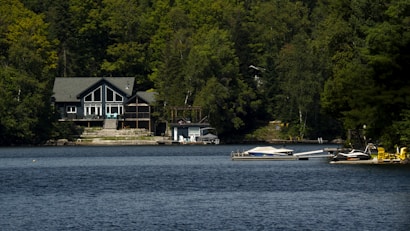 A serene lakeside scene featuring a large, modern house nestled among dense green trees. The calm, blue water reflects the greenery surrounding it. Several boats and docks are visible, including a small motorboat and a yellow watercraft. The setting suggests a peaceful, natural environment.