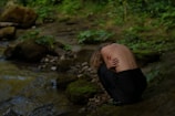 A person with blond hair and bare back crouches by a stream, surrounded by lush, green vegetation and moss-covered rocks. The environment is tranquil, and the individual appears to be in a contemplative or introspective state.