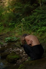 A person with long hair, partially unclothed, is crouched beside a rocky and moss-covered stream in a forest setting. The scene is enveloped in a sense of solitude and raw nature, with dense green foliage surrounding the area.