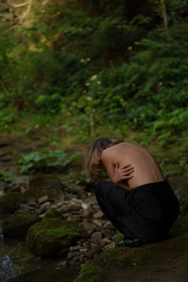 A person with long hair, partially unclothed, is crouched beside a rocky and moss-covered stream in a forest setting. The scene is enveloped in a sense of solitude and raw nature, with dense green foliage surrounding the area.