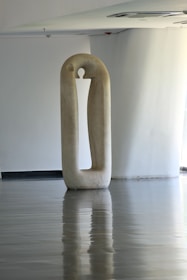 A tall, minimalist sculpture stands in a spacious, smooth-floored room. The sculpture, oval in shape with a cut-out center, has a reflective surface on the floor beneath. The environment is serene and modern, with soft natural lighting coming from an unseen source.