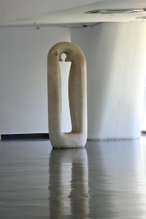 A tall, minimalist sculpture stands in a spacious, smooth-floored room. The sculpture, oval in shape with a cut-out center, has a reflective surface on the floor beneath. The environment is serene and modern, with soft natural lighting coming from an unseen source.