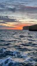 A dynamic ocean scene with rolling waves under a dramatic sky. The horizon features a sunset with vibrant orange and yellow hues, creating a stunning contrast against the dark silhouette of rocky cliffs.