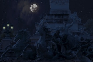 A moonlit scene featuring a dramatic sculpture of expressive horses and human figures. The focus is on the detailed forms of the statues against a dark night sky, with the full moon and stars adding a mystical element. Street lamps glow softly nearby, enhancing the ambiance.