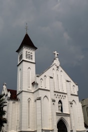 A large, white church with Gothic architectural features, including pointed arches and elaborate detailing. The building has a steeple with a cross on top and a small statue of a saint above the entrance. The sky is overcast, creating a somber atmosphere.