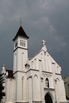 A large, white church with Gothic architectural features, including pointed arches and elaborate detailing. The building has a steeple with a cross on top and a small statue of a saint above the entrance. The sky is overcast, creating a somber atmosphere.