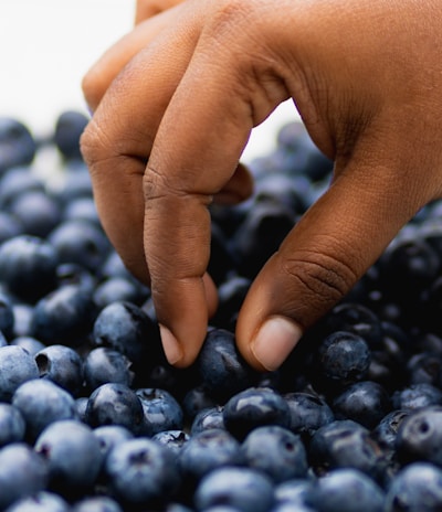 A close-up of a hand picking a blueberry from a pile. The hand appears to be carefully selecting the fruit, focusing on the texture and freshness of the blueberries. The image captures the smooth, deep blue surface of the berries in detail.