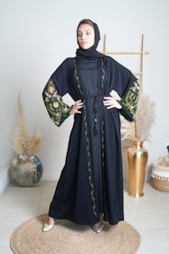 A woman stands confidently in an elegantly decorated room wearing a black abaya with intricate floral embroidery on the sleeves and trim. She is posed with her hands on her hips, conveying a sense of strength and poise. The background includes a decorative vase with pampas grass and a wooden frame, contributing to a sophisticated and minimalistic aesthetic.