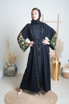 A woman stands confidently in an elegantly decorated room wearing a black abaya with intricate floral embroidery on the sleeves and trim. She is posed with her hands on her hips, conveying a sense of strength and poise. The background includes a decorative vase with pampas grass and a wooden frame, contributing to a sophisticated and minimalistic aesthetic.