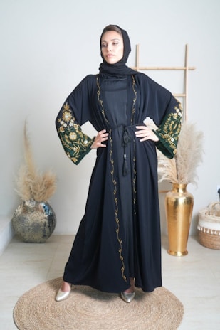 A woman stands confidently in an elegantly decorated room wearing a black abaya with intricate floral embroidery on the sleeves and trim. She is posed with her hands on her hips, conveying a sense of strength and poise. The background includes a decorative vase with pampas grass and a wooden frame, contributing to a sophisticated and minimalistic aesthetic.