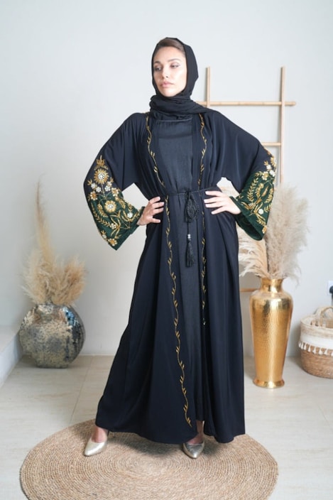 A graceful woman wearing a flowing black abaya with delicate embroidery standing in a sunlit garden.