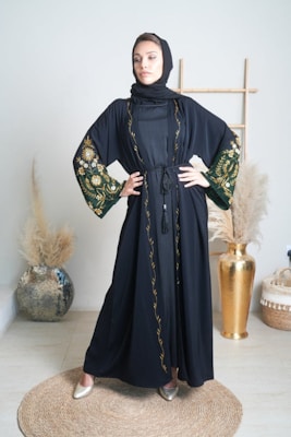A woman stands confidently in an elegantly decorated room wearing a black abaya with intricate floral embroidery on the sleeves and trim. She is posed with her hands on her hips, conveying a sense of strength and poise. The background includes a decorative vase with pampas grass and a wooden frame, contributing to a sophisticated and minimalistic aesthetic.