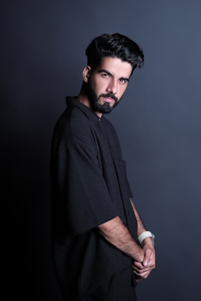 A bearded man with neatly styled hair is posing against a dark, plain background. He is wearing a loose-fitting black shirt and a white watch on his wrist. His gaze is directed towards the camera, and he has a composed expression.