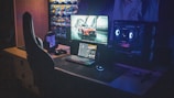 A dimly lit gaming setup with a large desk featuring a computer monitor displaying a sports car image. Below the monitor is a backlit keyboard and a glowing mouse. To the side is a gaming chair with a high back. The setup is illuminated by the RGB lighting from the computer tower on the right. Wall posters of cars decorate the background.
