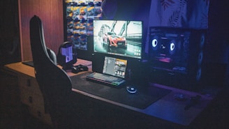 A dimly lit gaming setup with a large desk featuring a computer monitor displaying a sports car image. Below the monitor is a backlit keyboard and a glowing mouse. To the side is a gaming chair with a high back. The setup is illuminated by the RGB lighting from the computer tower on the right. Wall posters of cars decorate the background.