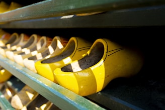 Bright yellow wooden clogs are lined up on a dark wooden shelf. The shoes are traditional in style, featuring curved vamps and are arranged neatly in rows, casting contrasting shadows.
