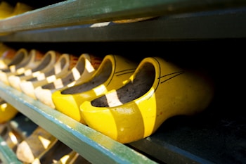 Bright yellow wooden clogs are lined up on a dark wooden shelf. The shoes are traditional in style, featuring curved vamps and are arranged neatly in rows, casting contrasting shadows.