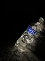 A large clear bottle labeled 'Absolut Vodka' is illuminated from within by a string of tiny white fairy lights. The bottle is placed on a fabric surface with a faint floral pattern, against a dark background, creating a cozy and artistic ambiance.