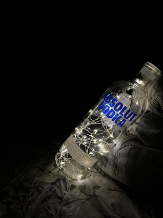 A large clear bottle labeled 'Absolut Vodka' is illuminated from within by a string of tiny white fairy lights. The bottle is placed on a fabric surface with a faint floral pattern, against a dark background, creating a cozy and artistic ambiance.