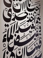 Large black Arabic script covers a white wall, featuring bold calligraphic letters with a variety of shapes and diacritical marks. The text is arranged in a visually dynamic and artistic manner, creating an intricate pattern.
