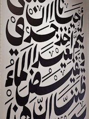 Large black Arabic script covers a white wall, featuring bold calligraphic letters with a variety of shapes and diacritical marks. The text is arranged in a visually dynamic and artistic manner, creating an intricate pattern.